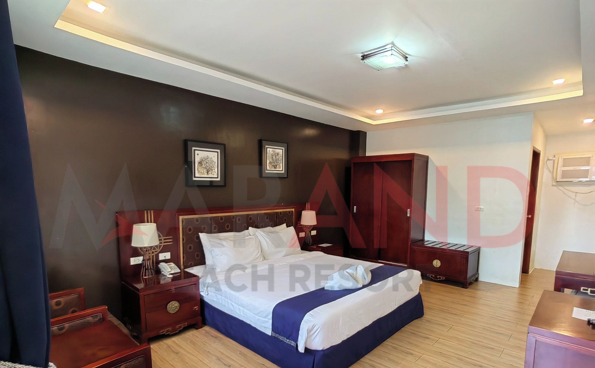 A Deluxe King bed in a hotel room with a dark brown wall, two framed pictures, a wooden wardrobe, and a bedside table with a lamp.