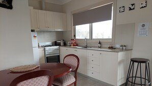 Une cuisine avec des meubles blancs, une cuisinière, un évier et une fenêtre donnant sur des arbres. On y trouve une table à manger avec des chaises et un tabouret.