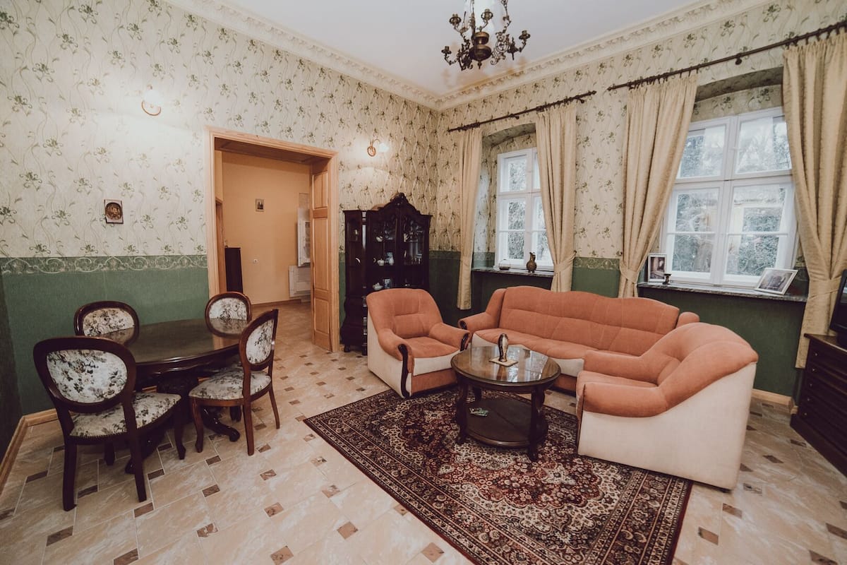 A traditional living room with a sofa set, a dining table with chairs, a patterned rug, and a bookshelf.