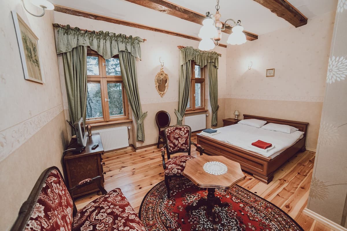 A cozy room with a wooden bed, a round table with a doily, two chairs, a desk with a computer, and two windows with curtains.