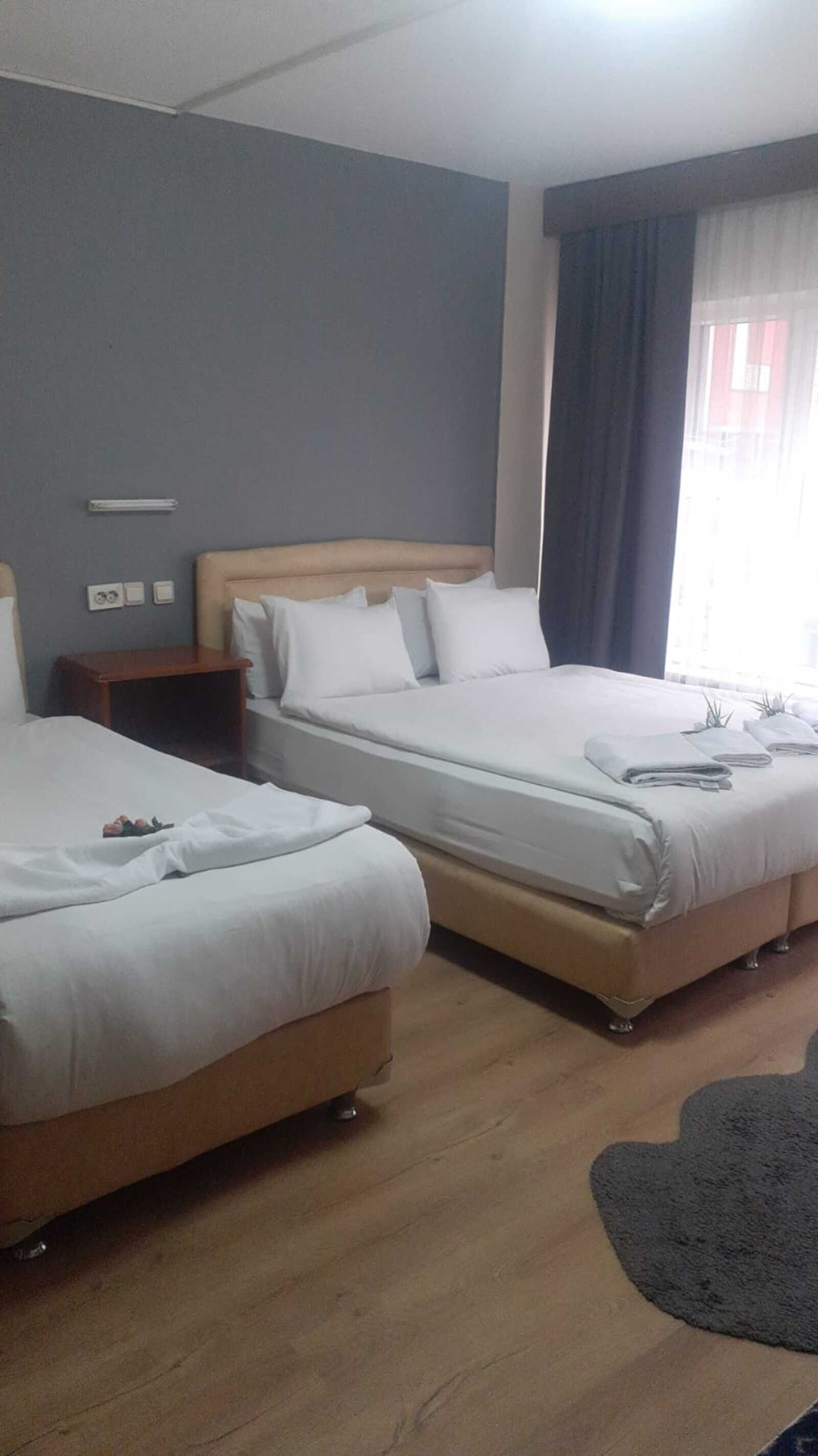 A hotel room with two beds, a wooden floor, a small bedside table, and a window with curtains.