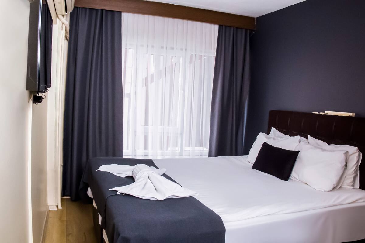 A hotel room with a large bed, dark curtains, and a window with sheer white curtains.