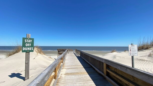A wooden boardwalk leading to a beach with a 'Stay Off Dunes' sign and a beach access sign with rules.