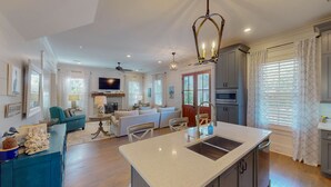 A spacious living room with a large sofa, a television mounted on the wall, and a fireplace. There is a kitchen area with a large island, a sink, and a microwave. The room has a ceiling fan and a chandelier.