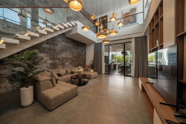 A spacious living area with a modern sofa, a coffee table, and a TV. A staircase with a glass railing leads to an upper level. - Tapa Ratan Uluwatu (Pecatu)
