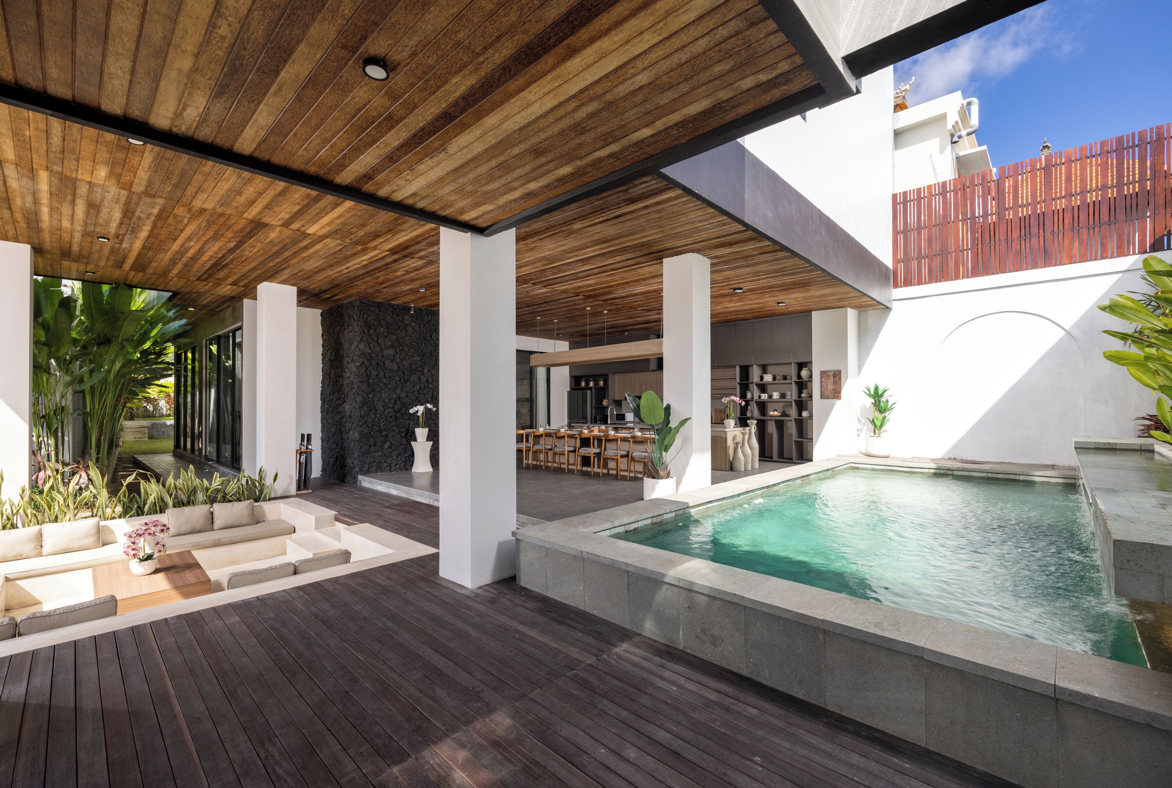 A modern outdoor pool area with a wooden deck, a stone wall, and a dining area.
