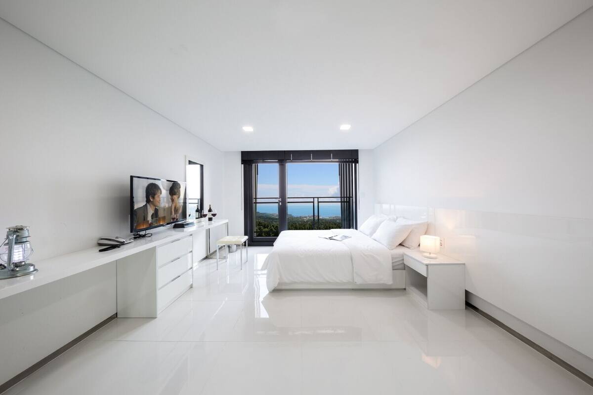 A modern bedroom with a large bed, a bedside table, a TV, and a window with a city view.
