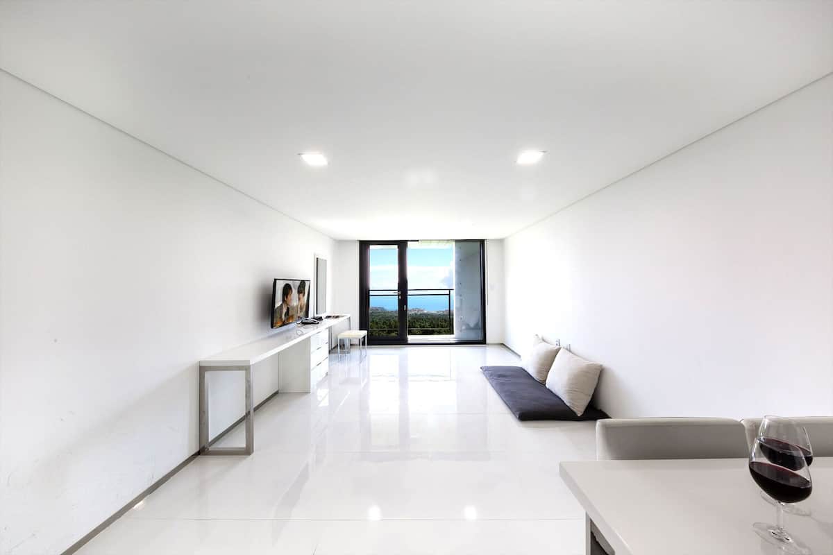 A modern, minimalist living room with a white color scheme, a large window offering a view of the outdoors, a flat-screen TV mounted on the wall, and a seating area with a sofa and a table.