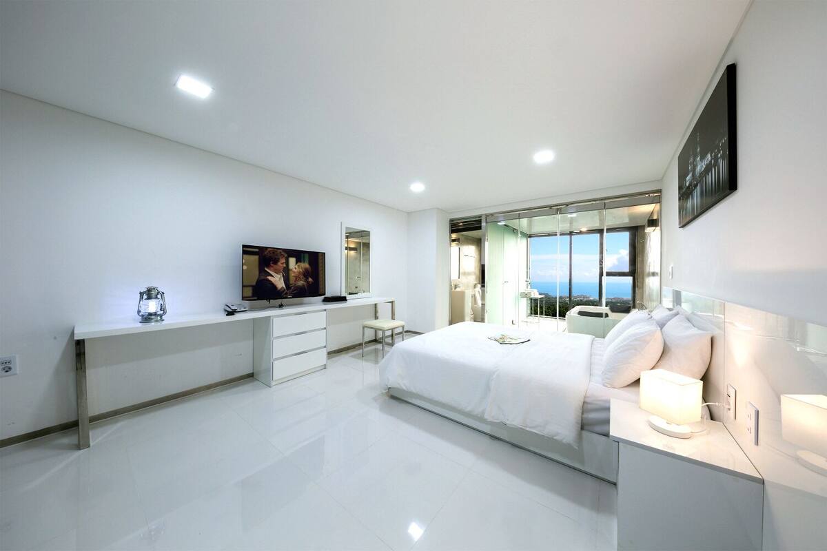 A modern hotel room with a large bed, a desk with a TV, a bathroom with a view, and a balcony.