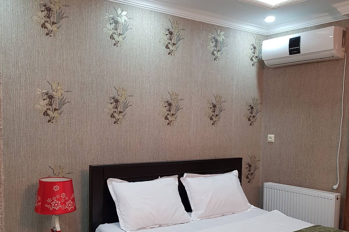 A neatly made bed with white bedding, a red lamp on a bedside table, and a patterned rug on the floor.
