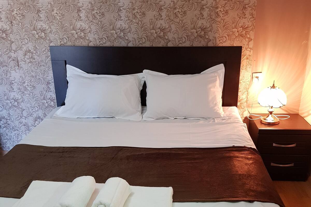 A neatly made bed with white linens, a brown headboard, and a bedside lamp.