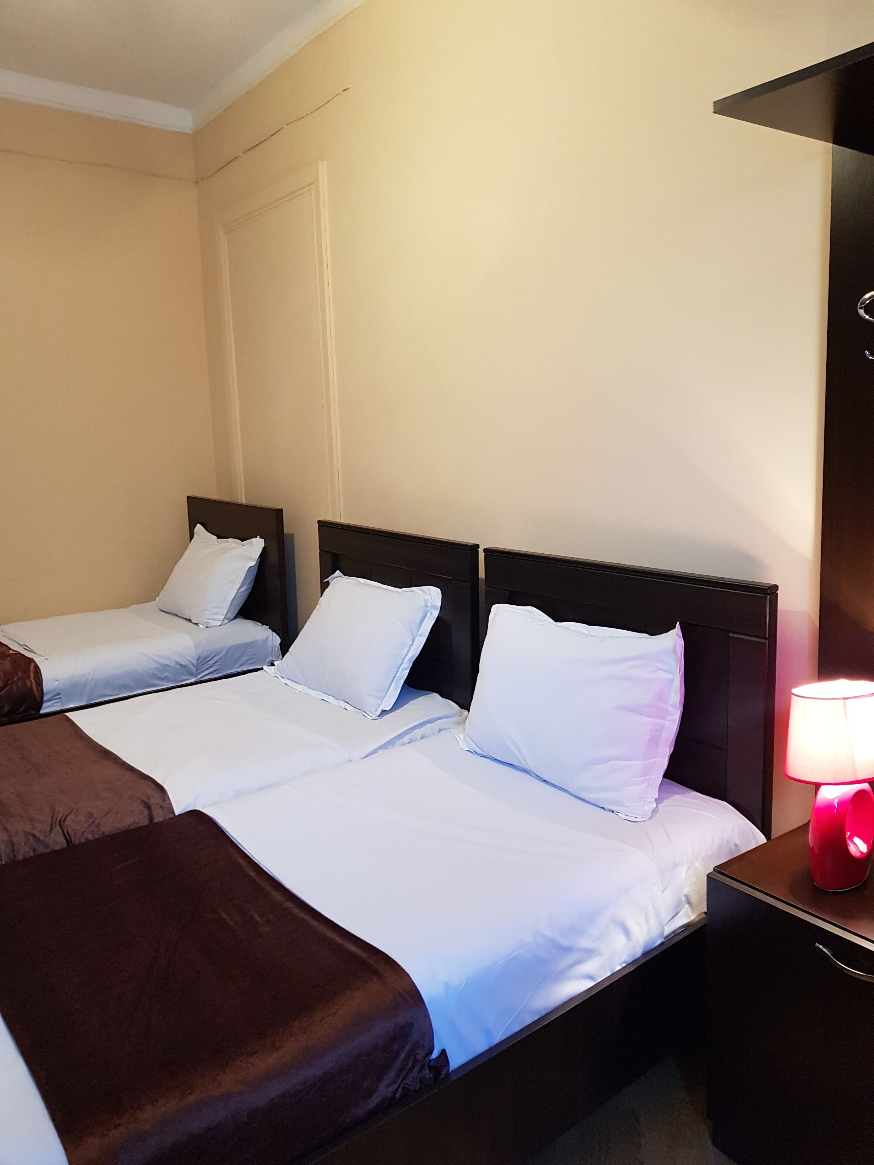 Una habitación de hotel con dos camas individuales, un banco marrón y una mesita de noche con una lámpara roja.