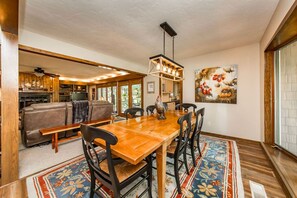 A dining area with a wooden table, black chairs, and a chandelier. There is a sofa and a fireplace in the background.