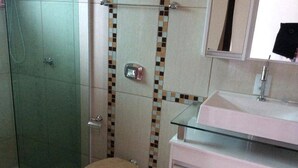 Een badkamer met een glazen douchecabine, toilet met een bloemrijk zitkussen, en een witte wastafel met een spiegel erboven.
