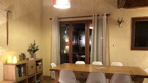A dining area with a wooden table and four white chairs, a wooden bookshelf, and a small plant.