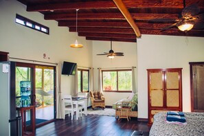 A spacious room with a wooden ceiling, a ceiling fan, a bed, a desk with a chair, a water dispenser, and large windows offering a view of greenery.