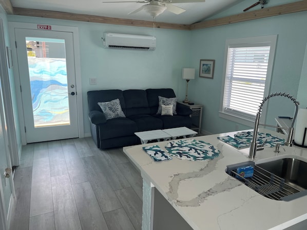 A compact living space with a sofa, a small table, a kitchenette with a sink, and a ceiling fan. - Bay Breeze - Cozy Beach Cottage across from the beach. Pet friendly! (Madeira Beach)