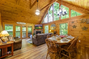 Relax in this cabin's living area, featuring a sofa, coffee table, and TV, with a dining space for meals and socializing, and large windows framing the surrounding greenery.