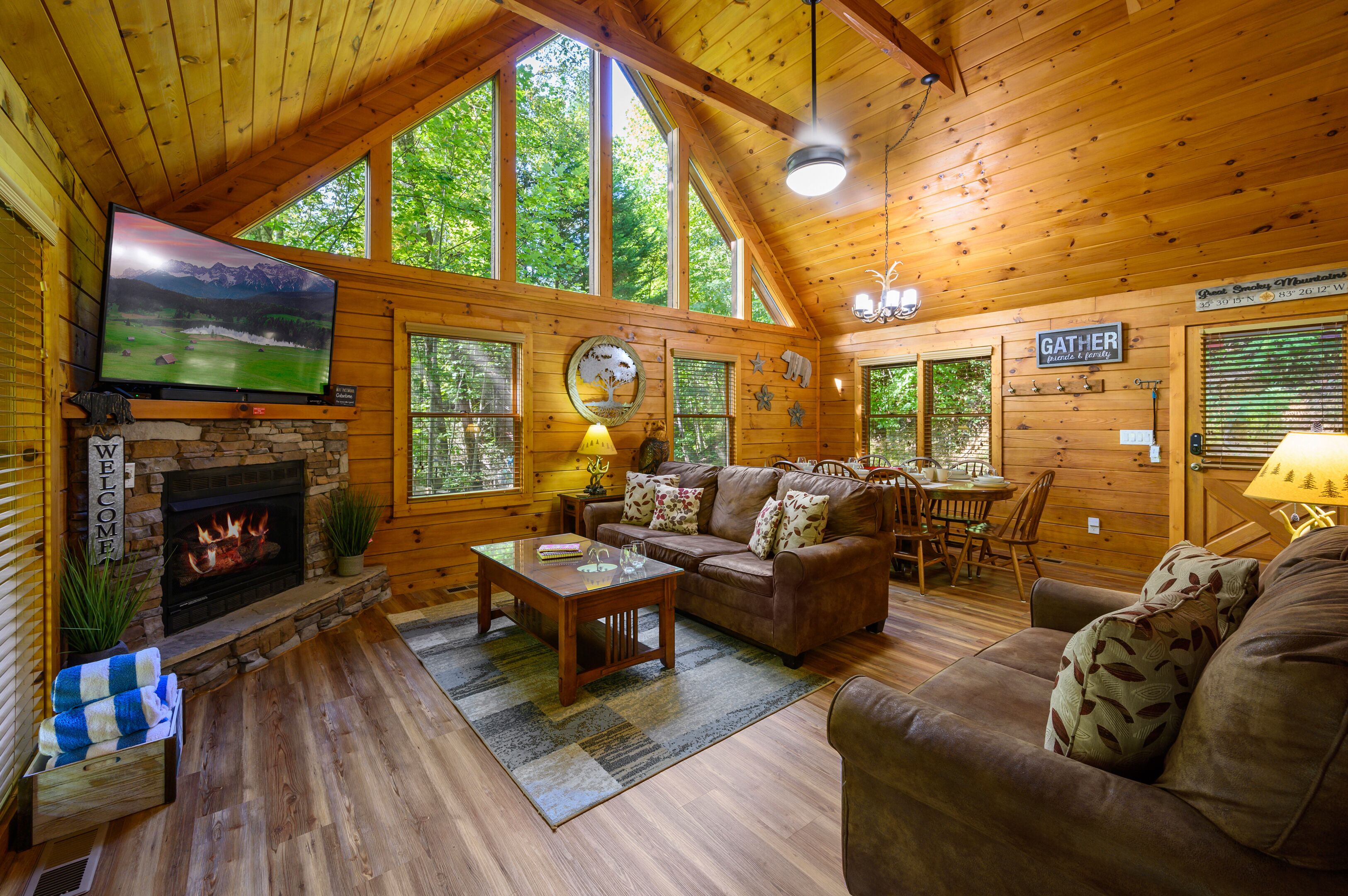 A cozy cabin interior with a stone fireplace, wooden furniture, and large windows offering a view of the forest.