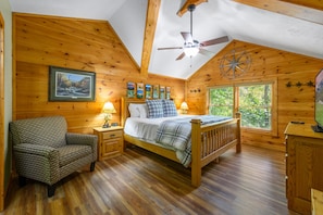Cozy bedroom with a wooden bed, nightstand, and armchair, featuring framed artwork and a window with a view of the surrounding greenery.