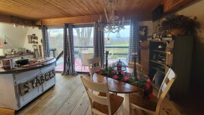 A rustic kitchen with wooden beams, a dining table with chairs, a chandelier, and a view of the outdoors through large windows. - Cozy Rustic Cabin In The Hamlet Of Shushan NY. with  incredible views (shushan)