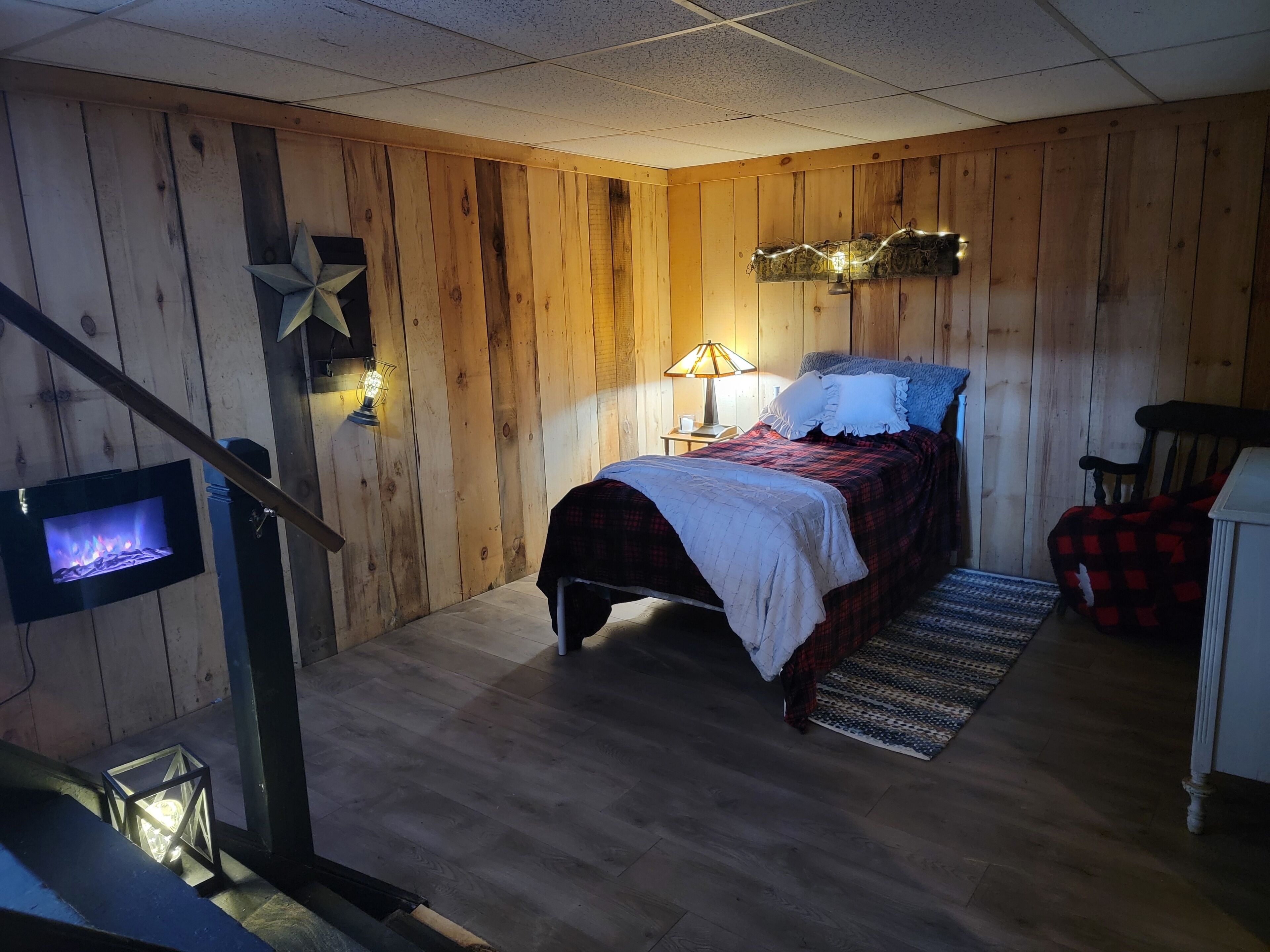 Een knus slaapkamer met een houten wand, een bed met een rood-wit geruit plaid, een nachtkastje met een lamp en een kleine decoratieve ster aan de wand.