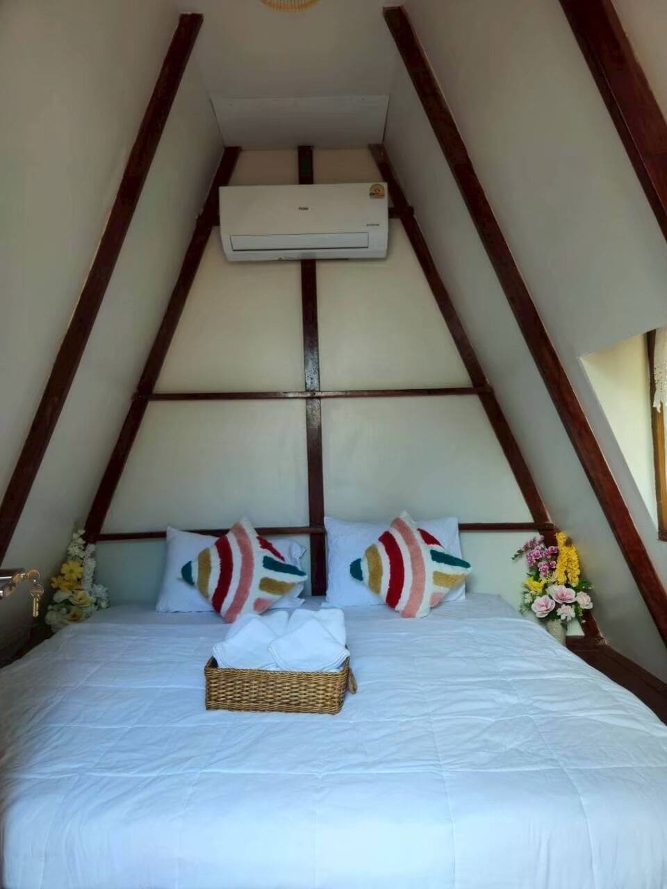 A room with a sloped ceiling, a bed with white bedding, two pillows, a wicker basket, and a wall-mounted air conditioner.