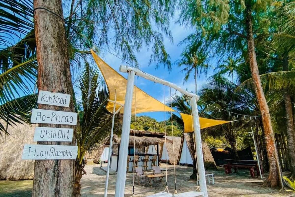 A beach area with a swing set, a thatched-roof hut, and a sign listing amenities like Koh Kood, Ao-Phrao, Chill Out, and I Lay Gramping.