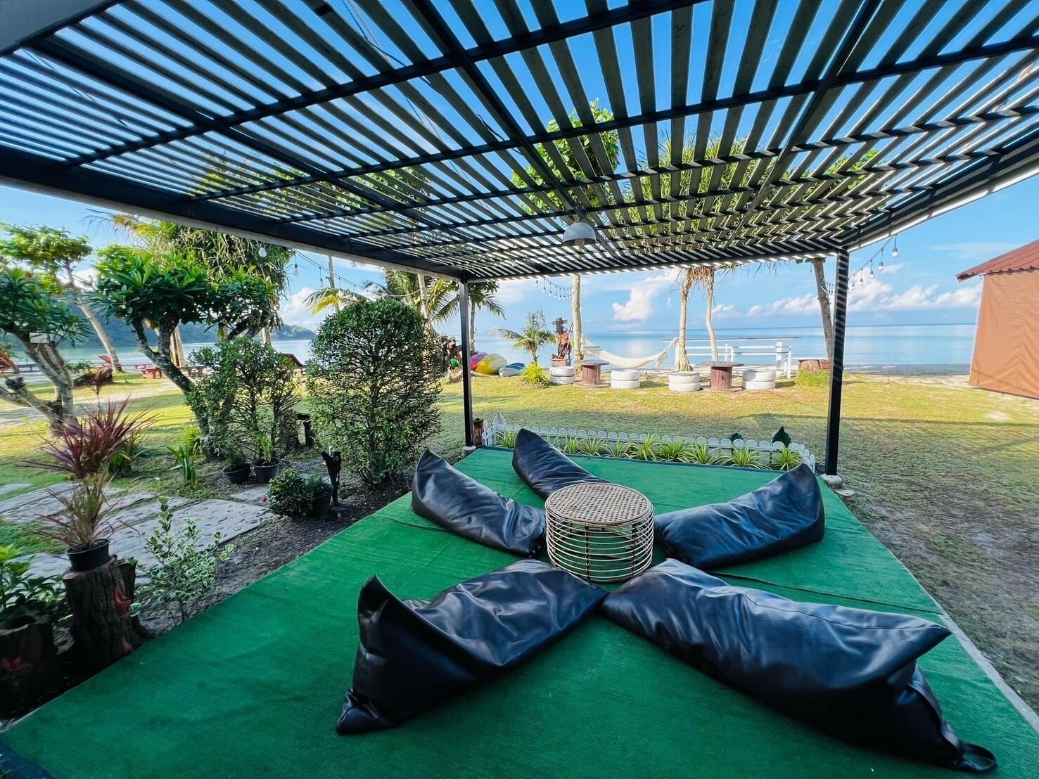A covered outdoor seating area with green cushions and a woven basket, overlooking a beach and palm trees.