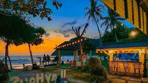 Un bar en la playa con mesas al aire libre, un letrero que dice "2UHY6I" y vista al océano al atardecer.