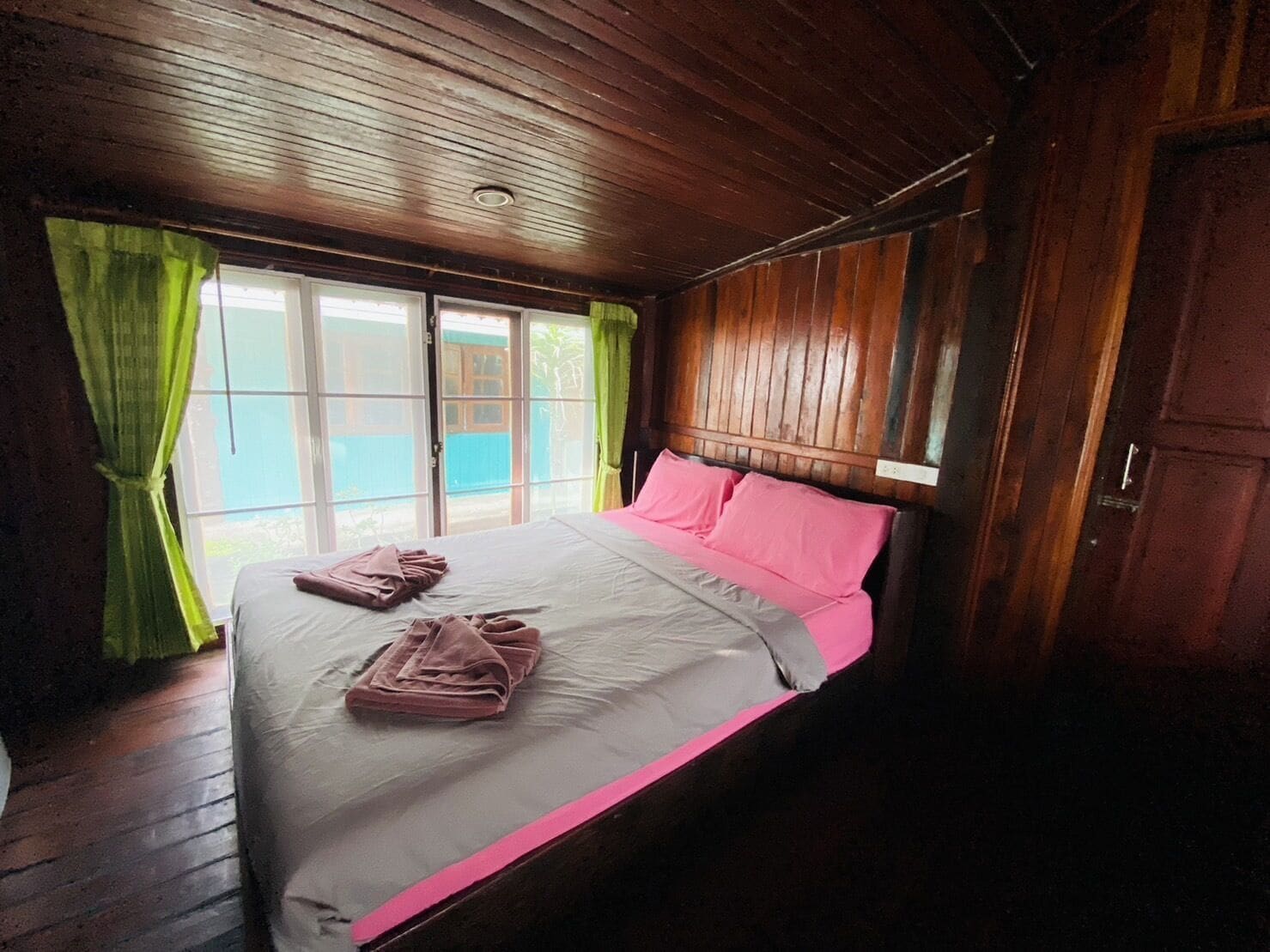 Een kamer met een houten plafond, een bed met roze en grijze beddengoed, en een raam met groene gordijnen.