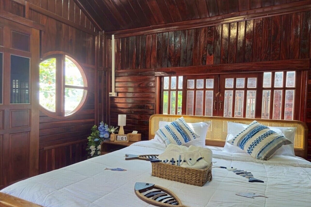 A wooden room with a bed, a round window, and a nightstand with a lamp.