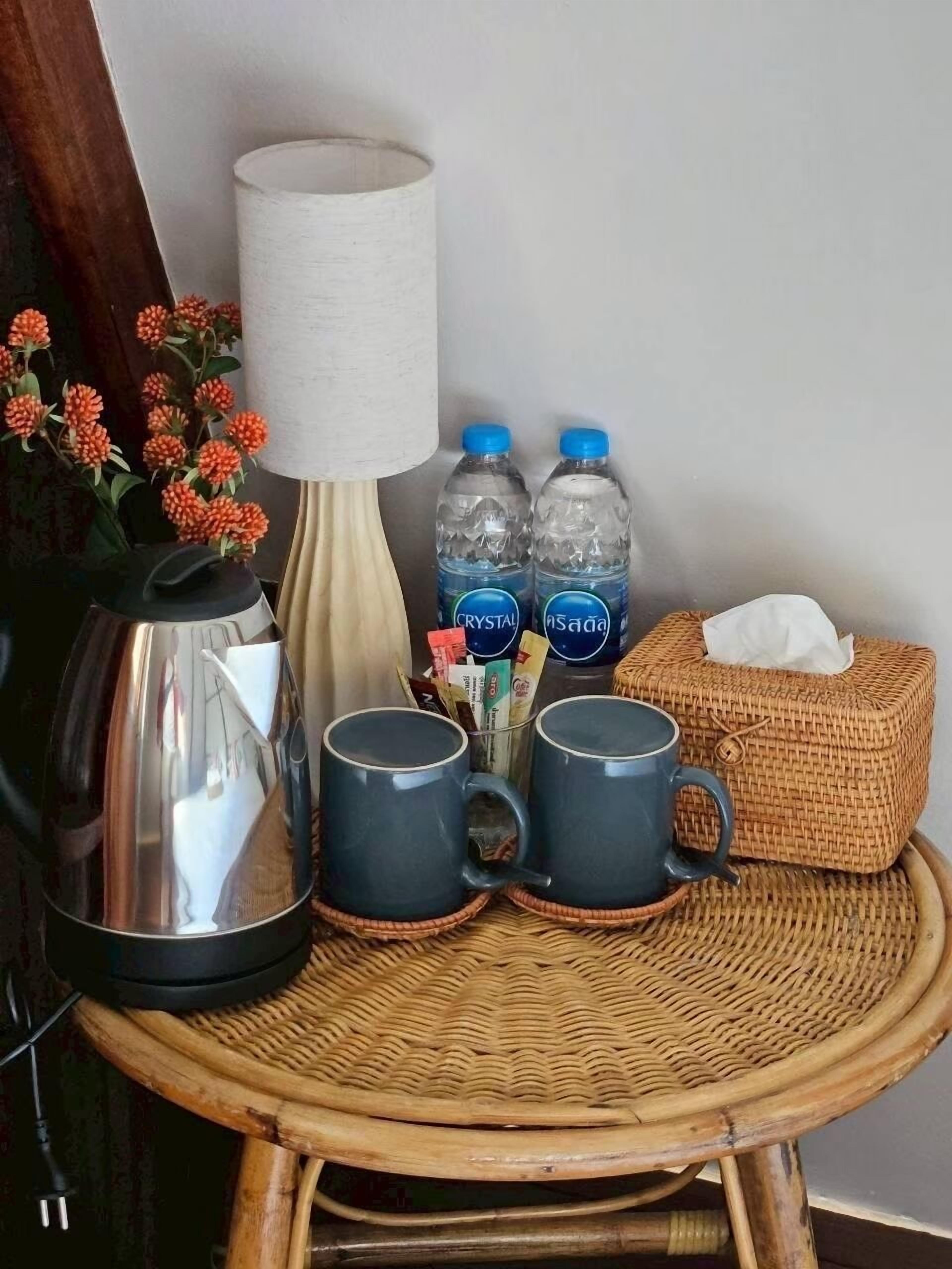 A wicker round table with a kettle, two mugs, two water bottles, a tissue box, and a vase with orange flowers.