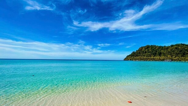 Una playa con agua turquesa cristalina, una orilla arenosa y una colina boscosa al fondo.