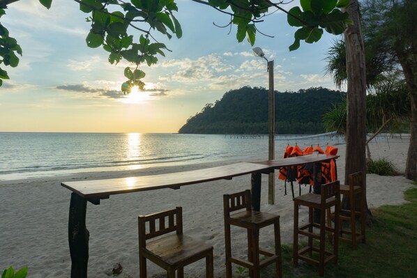 A wooden table and chairs on a sandy beach with a view of the ocean and a forested hill in the background. - I-Lay House Koh Kood (Ko Kood)