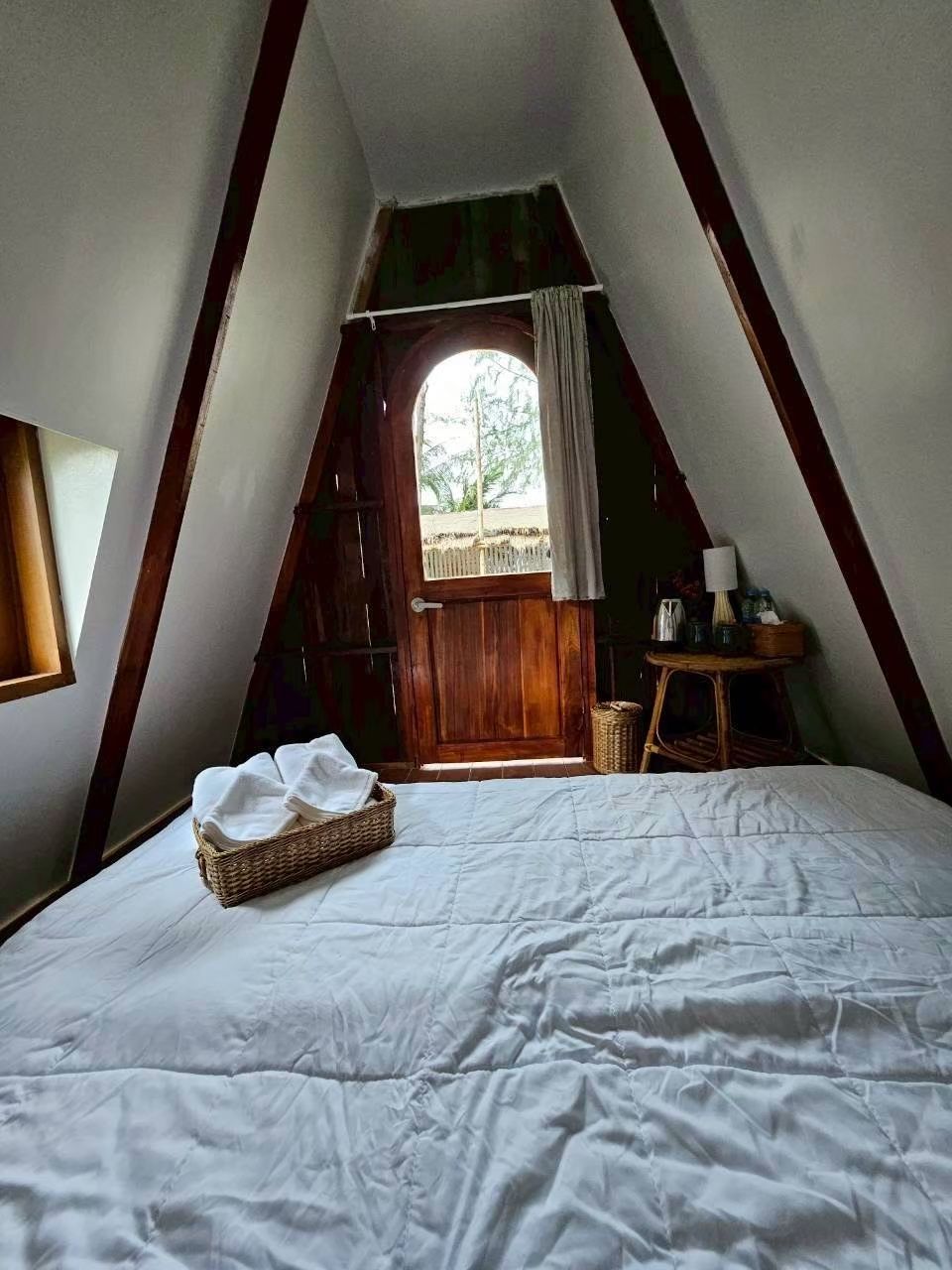 A bedroom with a large bed, a wooden door, a small table with a lamp, and a window with curtains.