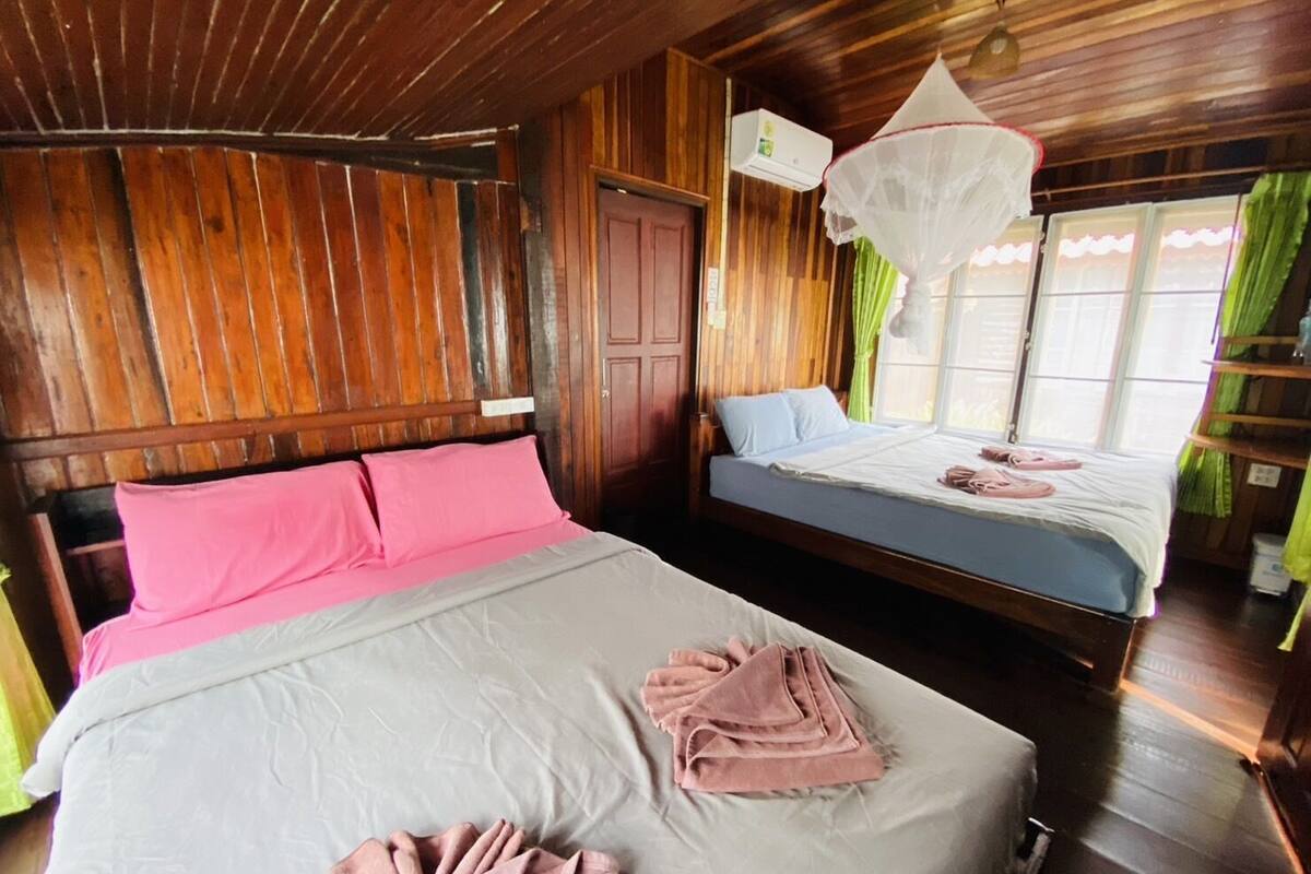 A room with two beds, wooden walls, a ceiling fan, and a window with curtains.