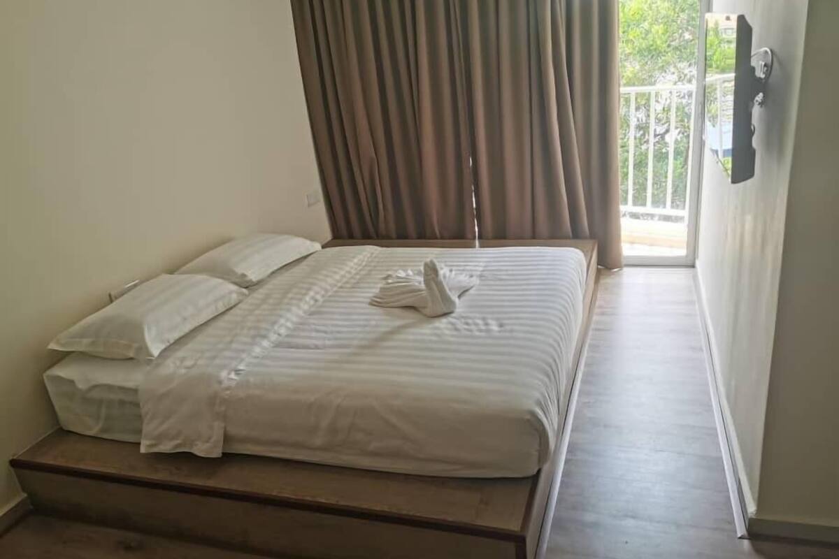 A single bed with a wooden frame, a white bedspread, and two pillows. There is a window with curtains, and a door with a handle is visible.