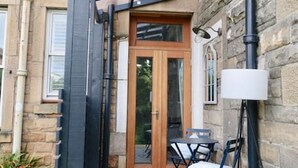Een stenen gebouw met een houten deur, een klein terras met een tafel en stoelen, en een lantaarnpaal.