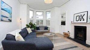 Een moderne woonkamer met een donkergrijze bank, een ronde salontafel en een open haard. Er is een groot raam met zonwering, een schilderij aan de muur en een bordje met de letter 'OK'.