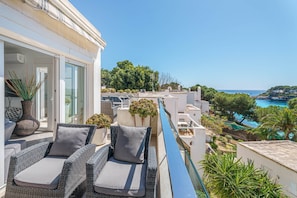 A balcony with wicker furniture and potted plants, overlooking a coastal area with trees and a clear sky.