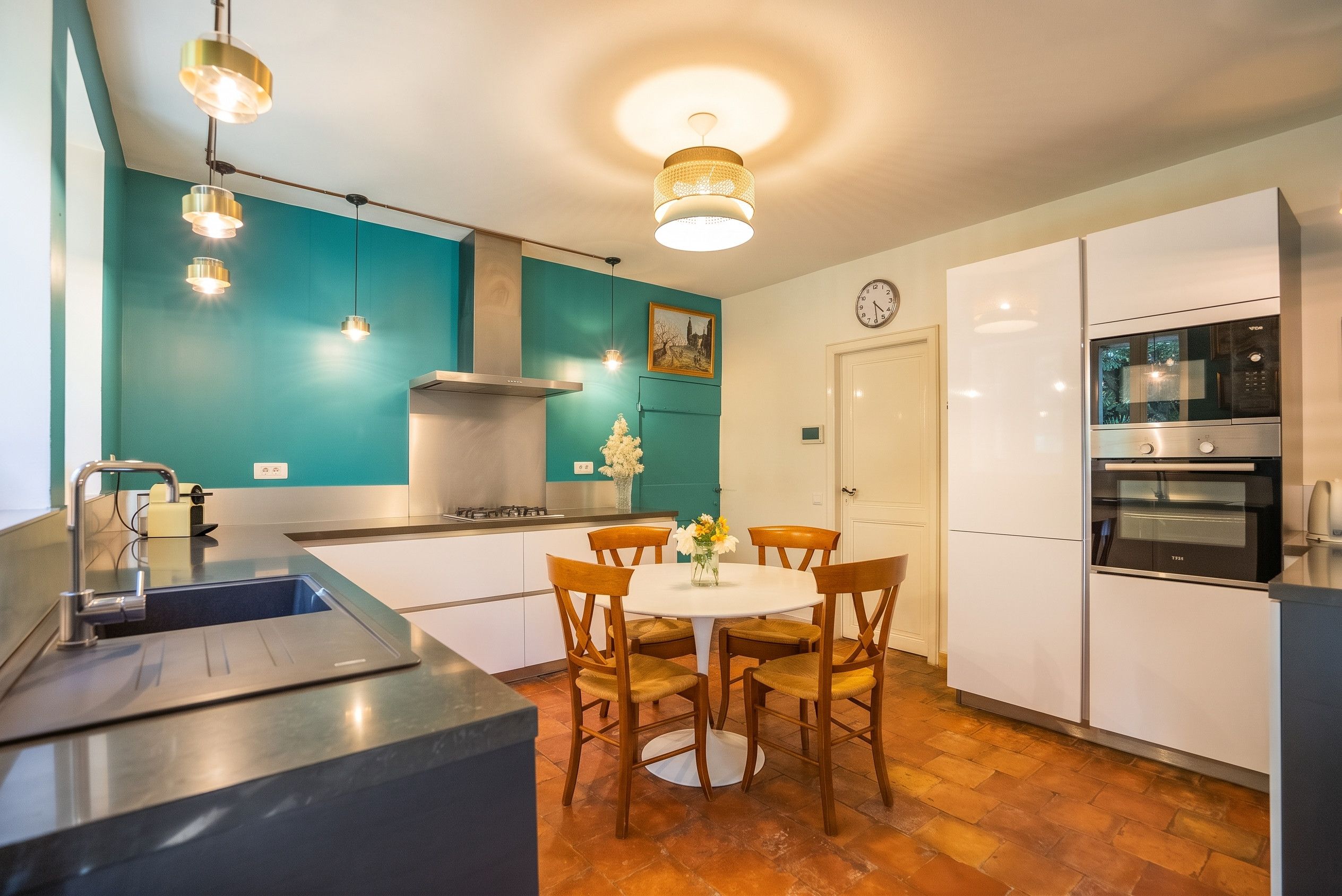 A modern kitchen with a central island, white cabinets, teal walls, and a dining area with wooden chairs.