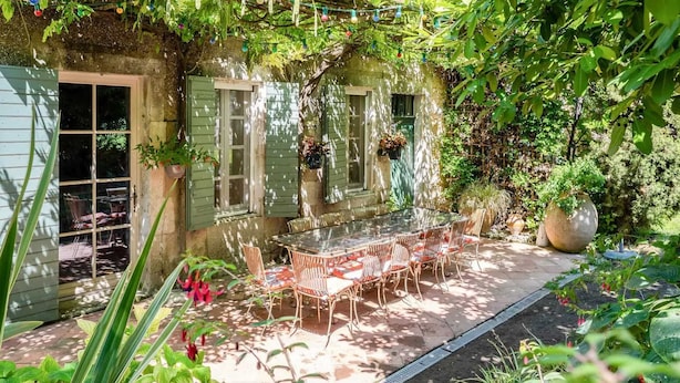 Une agréable terrasse extérieure, dotée d’une table à plateau de verre et de chaises en osier, entourée d’une végétation luxuriante et de plantes en pot.