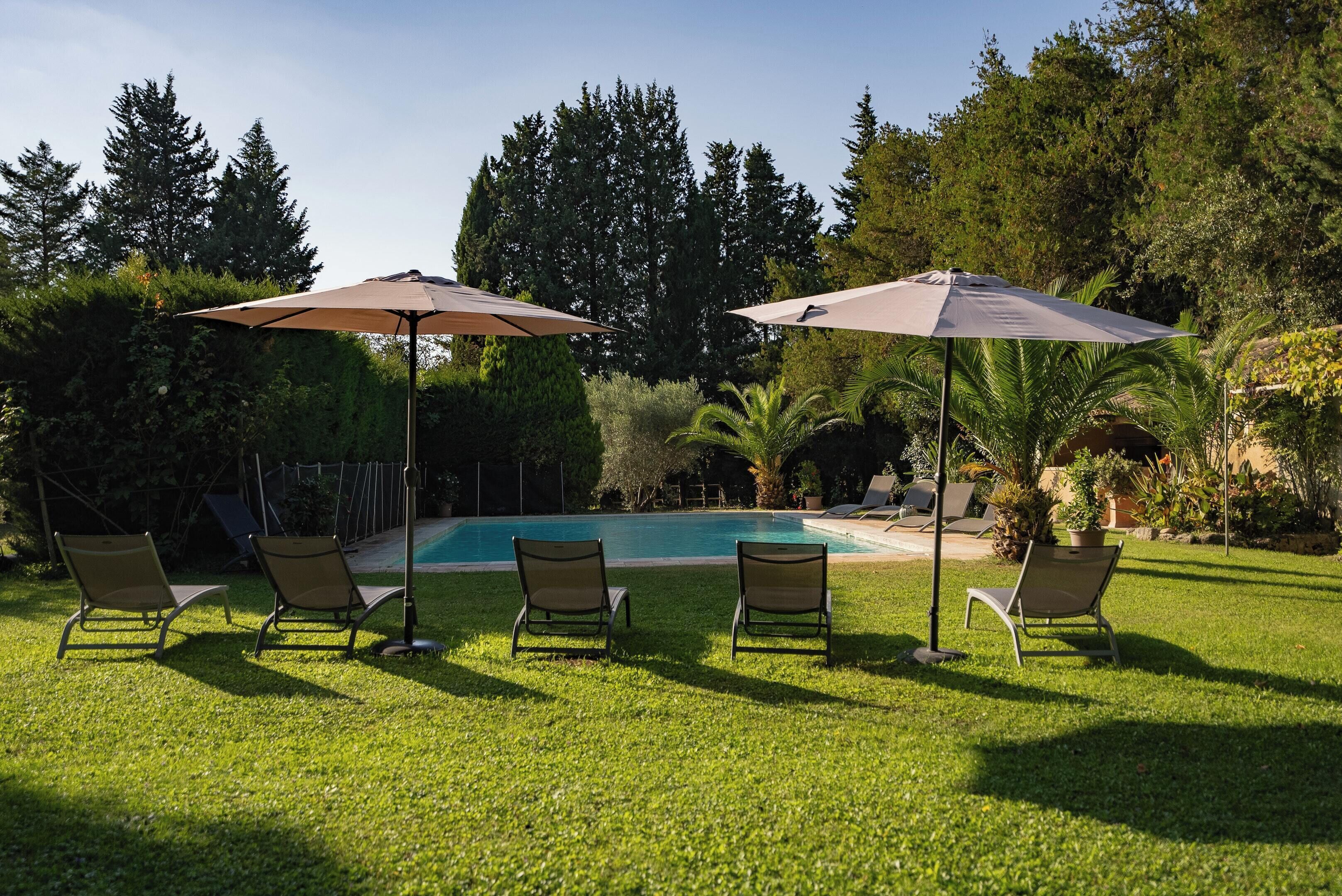 Un espace aménagé au bord de la piscine, comprenant quatre chaises longues et deux parasols, le tout entouré d’une végétation luxuriante et d’arbres.