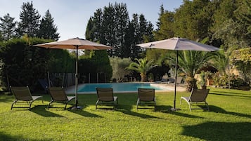 Un espace aménagé au bord de la piscine, comprenant quatre chaises longues et deux parasols, le tout entouré d’une végétation luxuriante et d’arbres.