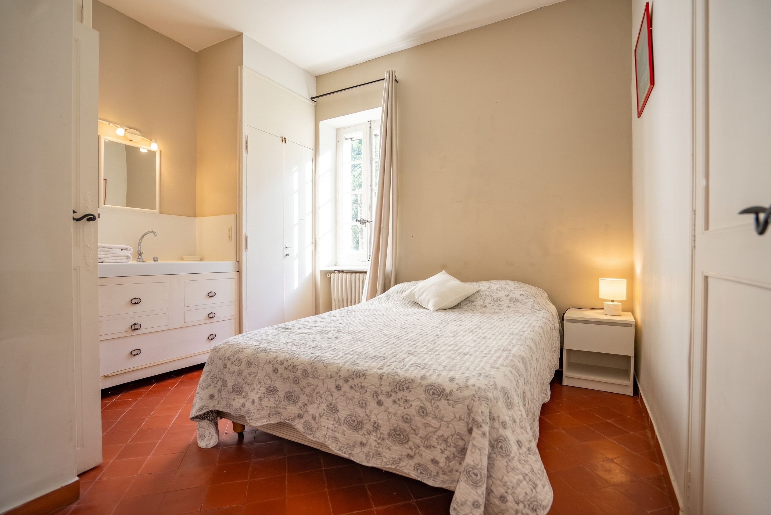 Ein Schlafzimmer mit einem Bett, einem Nachttisch mit Lampe, einer Kommode mit Spiegel und einem Fenster mit Vorhängen.