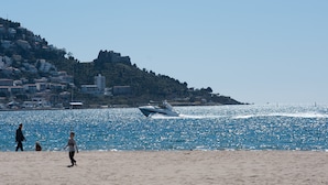 Une plage avec un bateau sur l’eau, des personnes qui se promènent et des bâtiments sur une colline en arrière-plan.