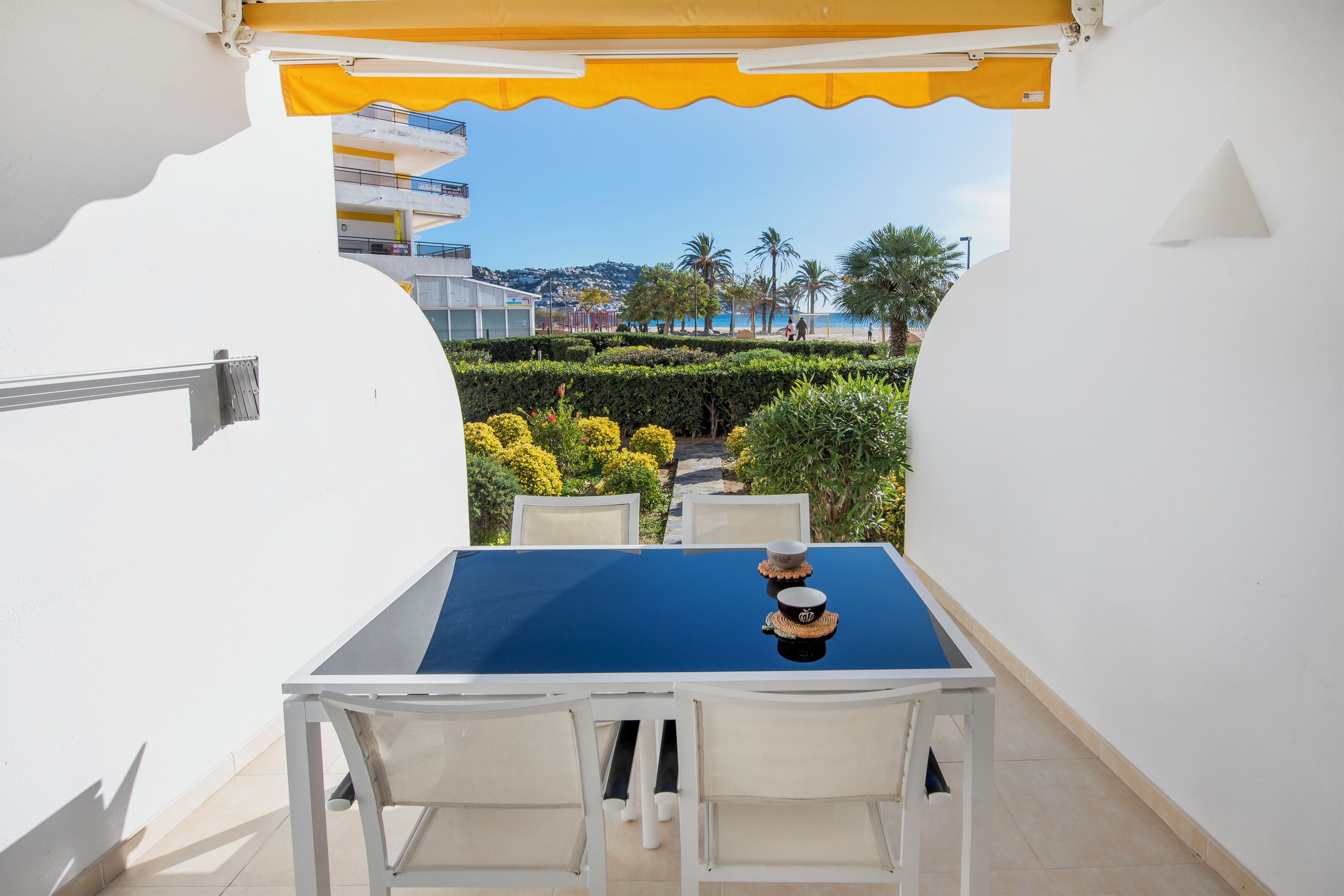 A table with a blue tablecloth and two chairs, a yellow awning, and a view of a landscaped garden and buildings in the background.