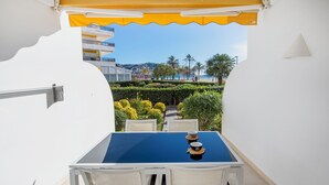 Une table recouverte d’une nappe bleue, deux chaises, un auvent jaune et, en arrière-plan, une vue sur un jardin aménagé et des bâtiments.