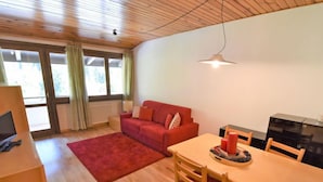 Un salon chaleureux avec un canapé rouge, une table à manger en bois et des chaises. On y trouve un téléviseur posé sur un meuble en bois, un radiateur et une fenêtre avec des rideaux.